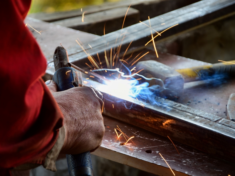 A person in a red sleeve uses a welding tool on a metal frame during local metal fabrication, producing a bright arc and flying sparks.