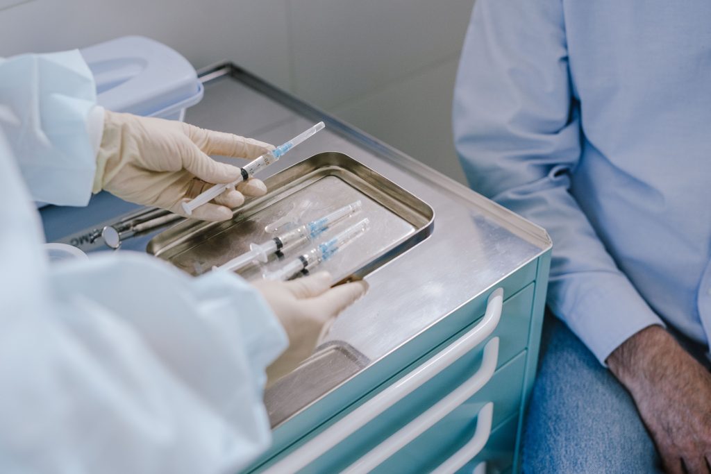 A medical professional in gloves holds a syringe over a stainless steel tray containing additional syringes next to a local patient.