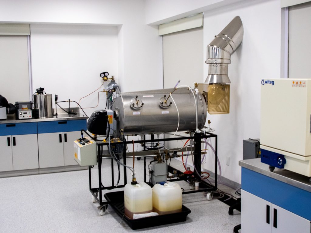 A Wisconsin laboratory setting featuring a large horizontal stainless steel reactor vessel on a wheeled stand, connected to a metal exhaust vent and two plastic containers of liquid on a drip tray for local fabrication.